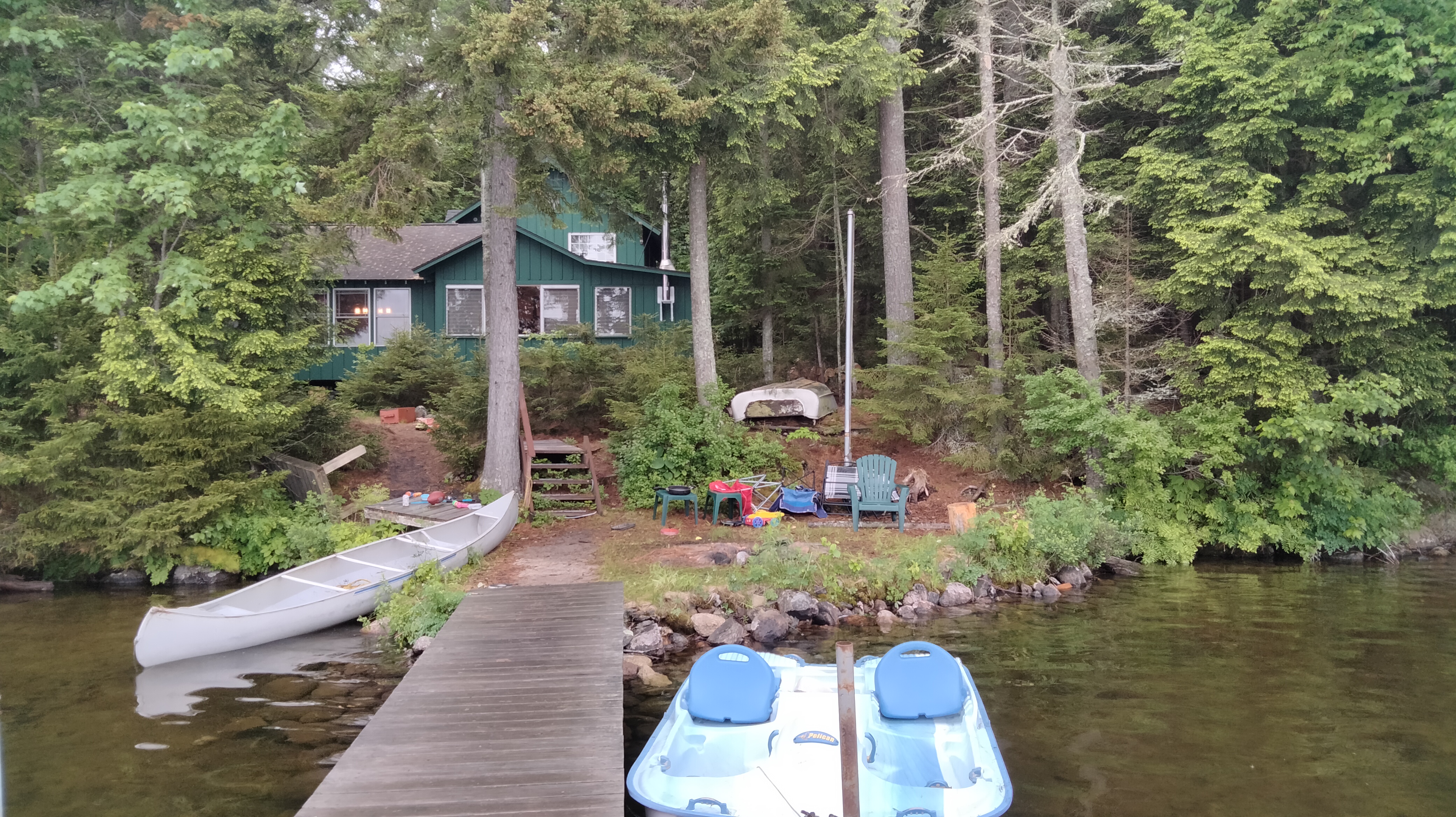 A green cabin in the woods. A light is on in the large living room window.