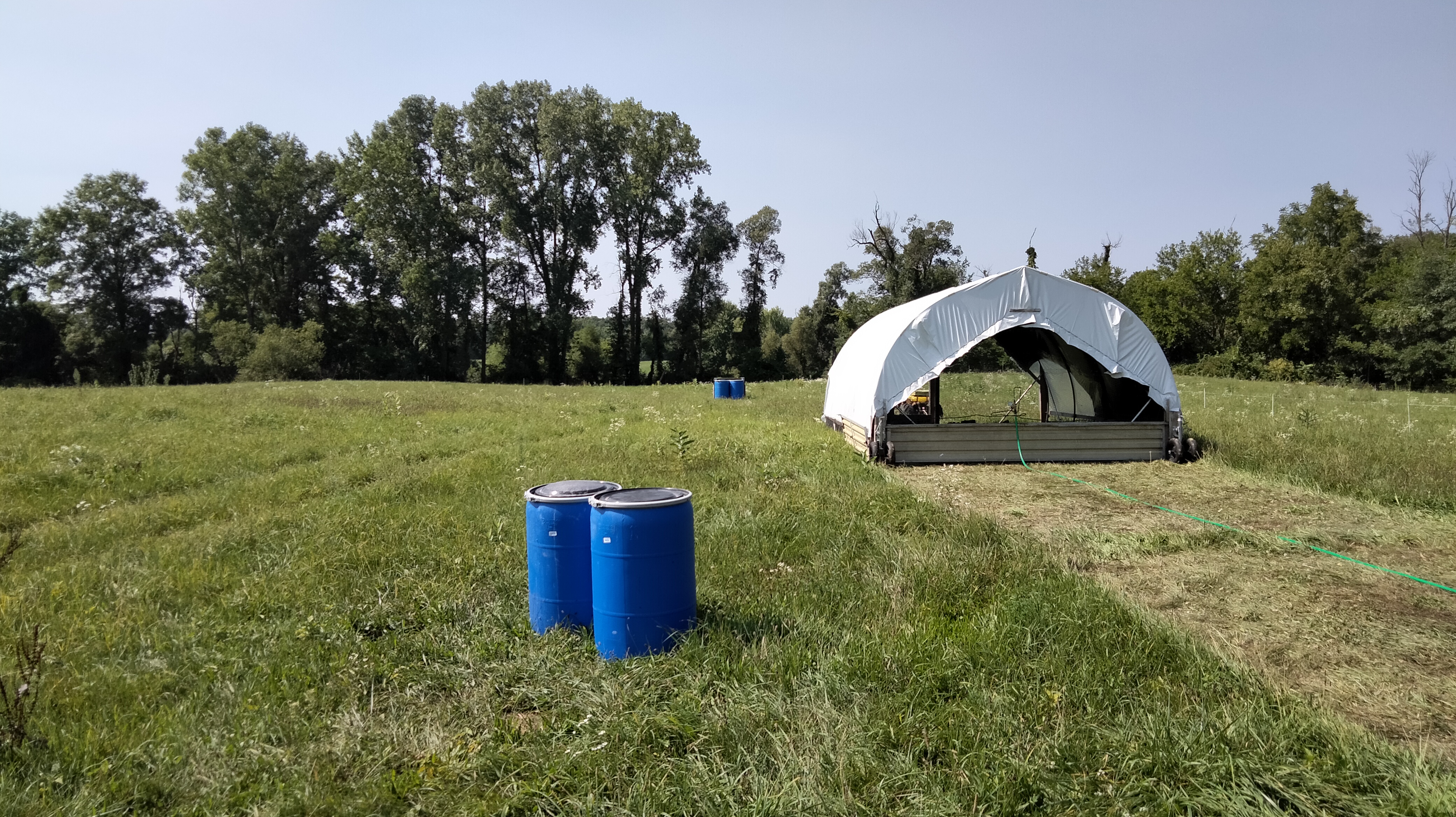 A white trailer in a field. Blue food barrels are in the foreground.