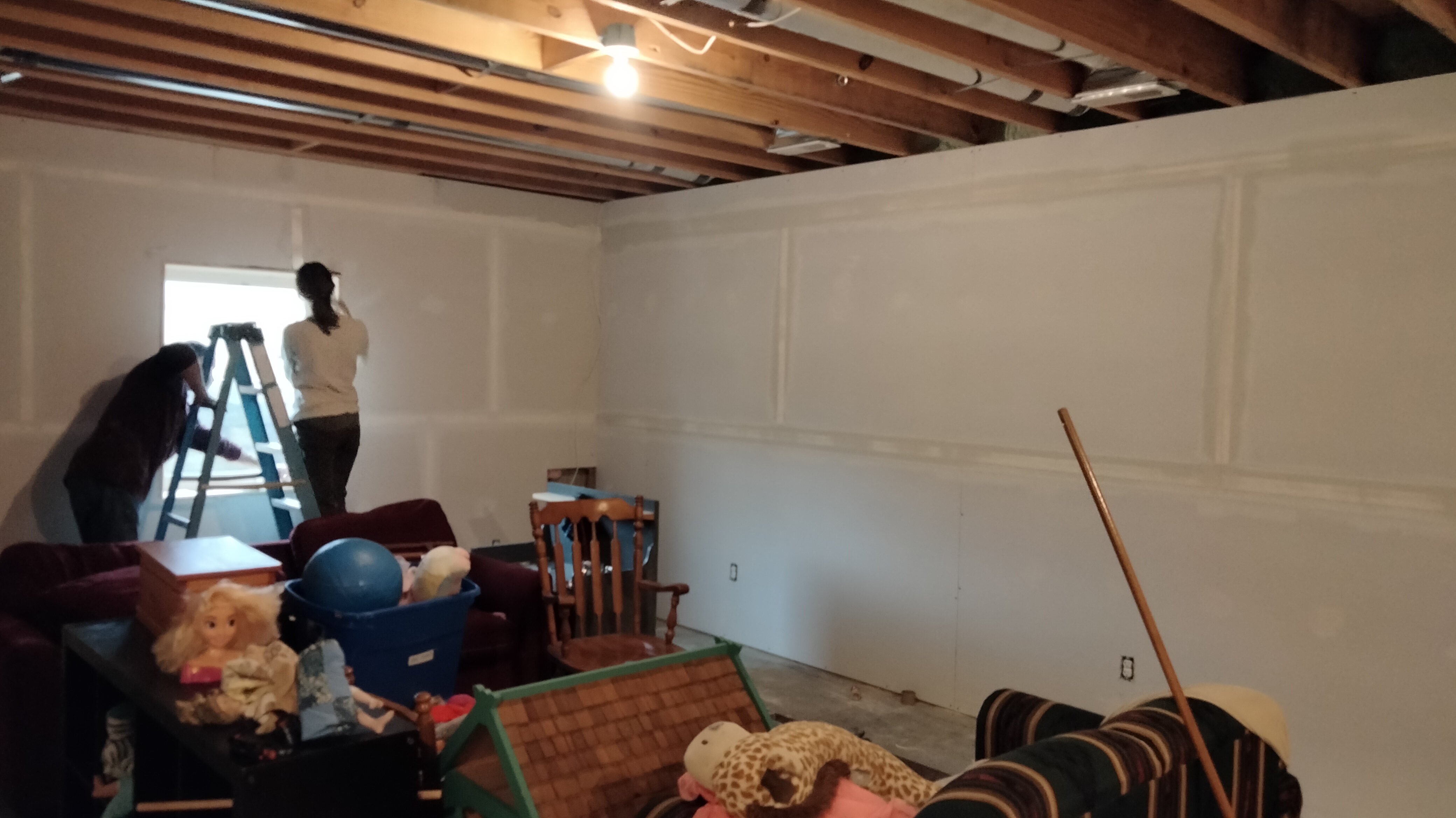 Two people, a man and a woman, are plastering drywall seams in a partially finished basement.