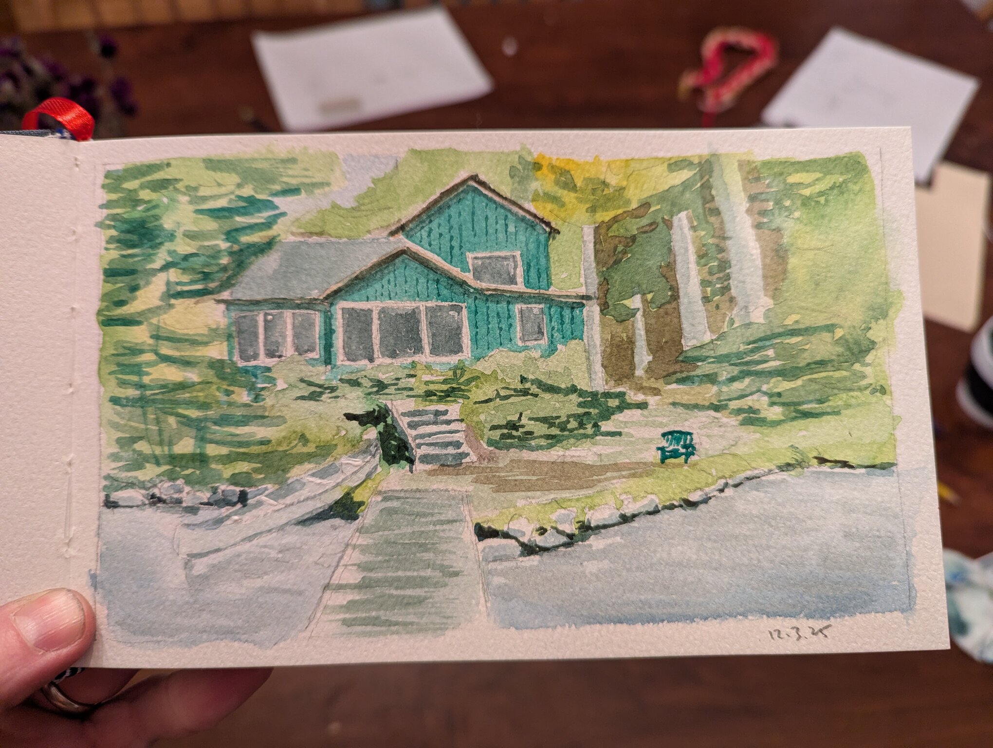 A green house set deep back into the woods. The viewer's perspective is looking at the house from a dock on the lake.