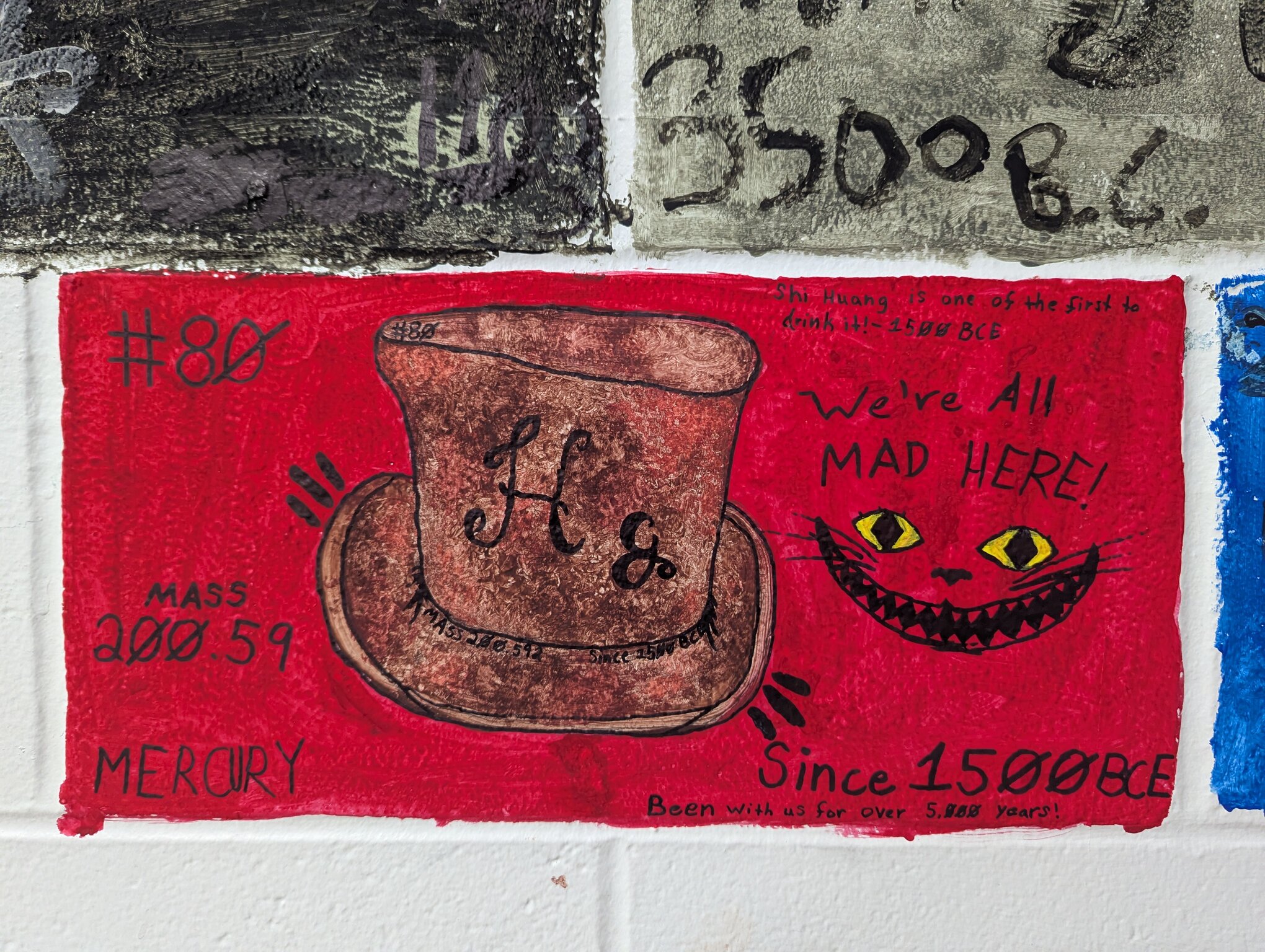 A painted cinderblock. A large brown top hat has the symbol for mercury, Hg, painted in the middle. It says, "We're all mad here" with the Cheshire Cat's face because mercury was used in felting and the workers often went insane due to mercury poisoning.