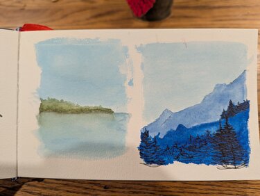 Two small landscapes. The left is a peninsula of trees in the distance with water in the foreground. The trees are lightly reflected in the water. The right is a mountainscape in all blues. A light blue sky is above pale blue mountains which are covered by a much darker blue range in the foreground. Black pen evergreen trees are also in the foregrouns.